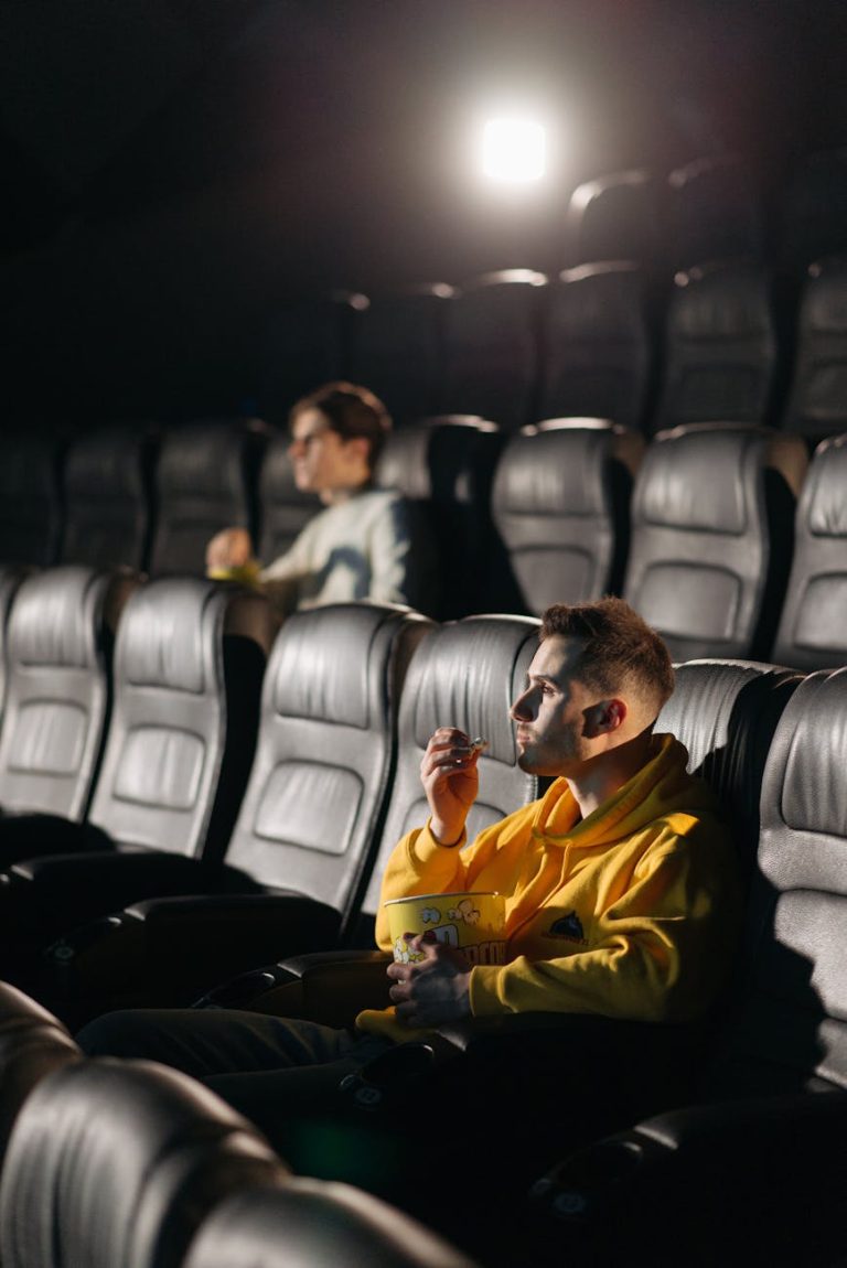 A man in a yellow hoodie eating popcorn alone in a dimly lit movie theater.