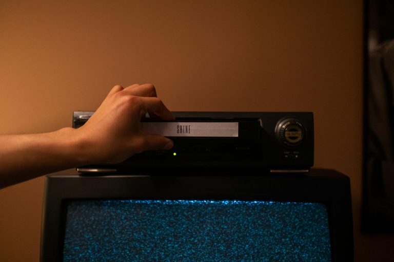 A hand inserting a VHS tape labeled 'SHINE' into a vintage player. Nostalgic home movie setup.